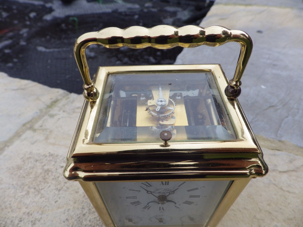 A polished brass LEpee carriage clock featuring bevelled glass panels, a white dial with Roman numerals, a moon phase aperture, and three subsidiary calendar dials. View II