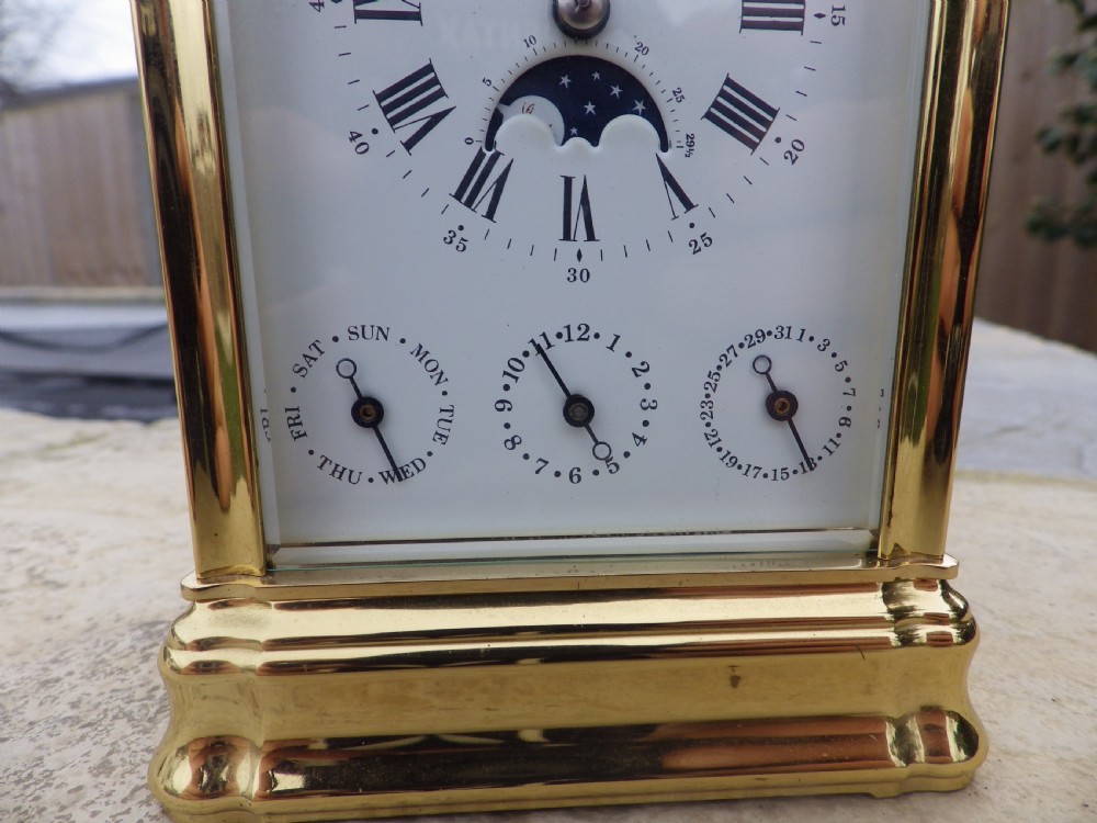 A polished brass LEpee carriage clock featuring bevelled glass panels, a white dial with Roman numerals, a moon phase aperture, and three subsidiary calendar dials. View III