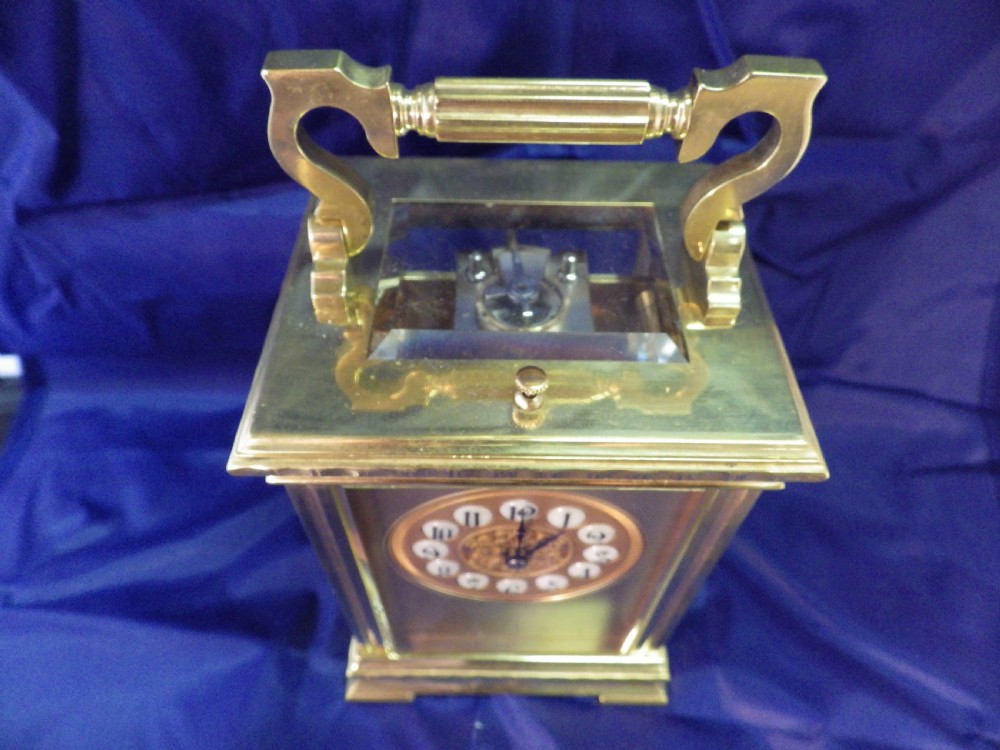 An antique brass French repeating carriage clock featuring an ornate circular dial with enamel numeral cartouches and a top carrying handle. View VII