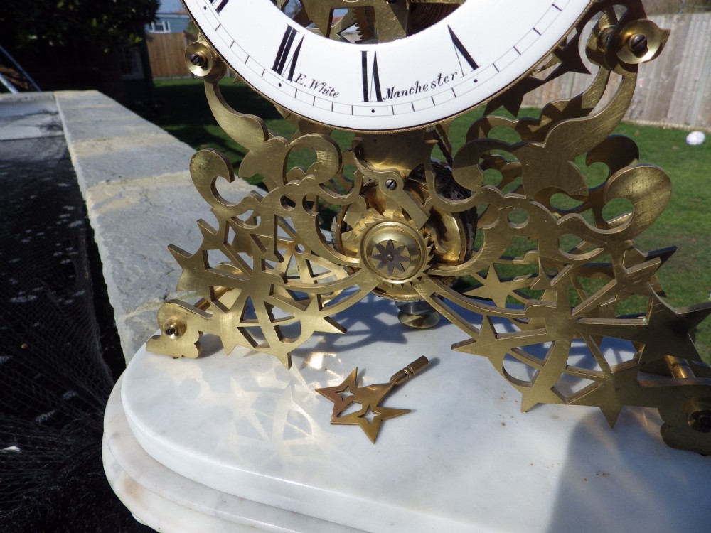 An antique Victorian brass skeleton clock featuring exposed internal mechanical gearing, a top-mounted bell, an enamel chapter ring with Roman numerals, and a white marble base. View II