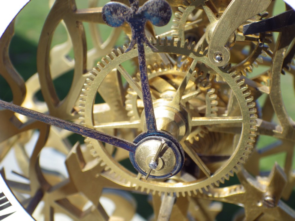 An antique Victorian brass skeleton clock featuring exposed internal mechanical gearing, a top-mounted bell, an enamel chapter ring with Roman numerals, and a white marble base. View IV
