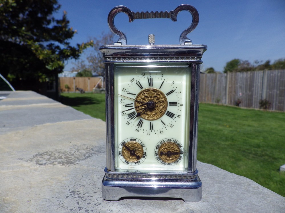 A silver-plated French carriage clock featuring a top carrying handle, a main white enamel dial with Roman numerals, and two subsidiary calendar dials set within a rectangular glass-panelled case.