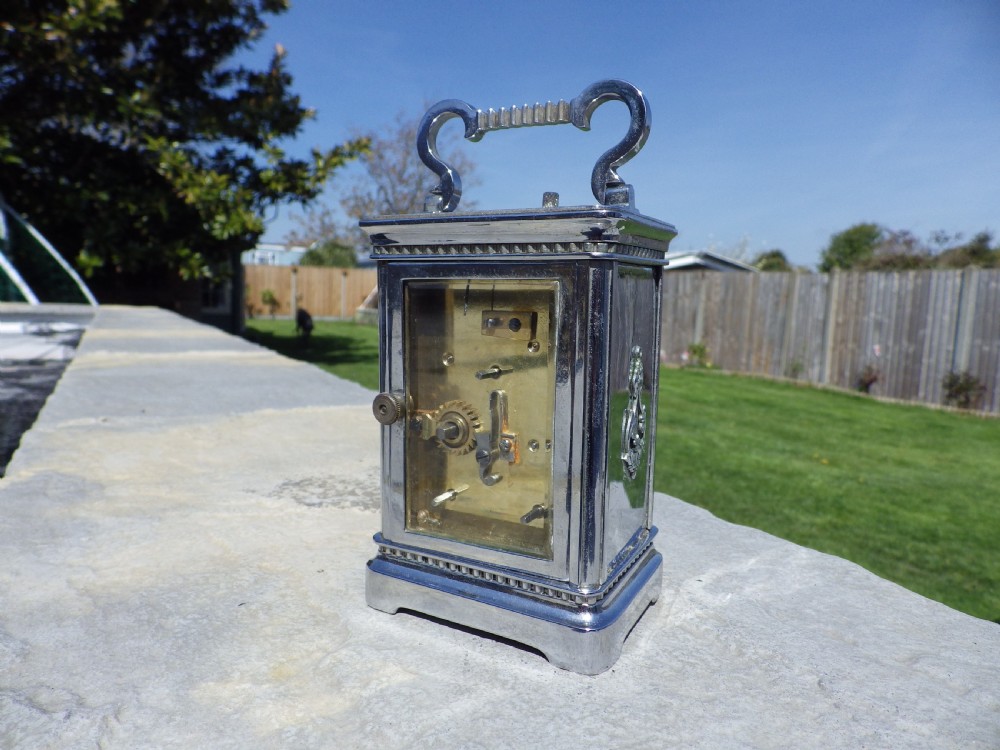 A silver-plated French carriage clock featuring a top carrying handle, a main white enamel dial with Roman numerals, and two subsidiary calendar dials set within a rectangular glass-panelled case. View III