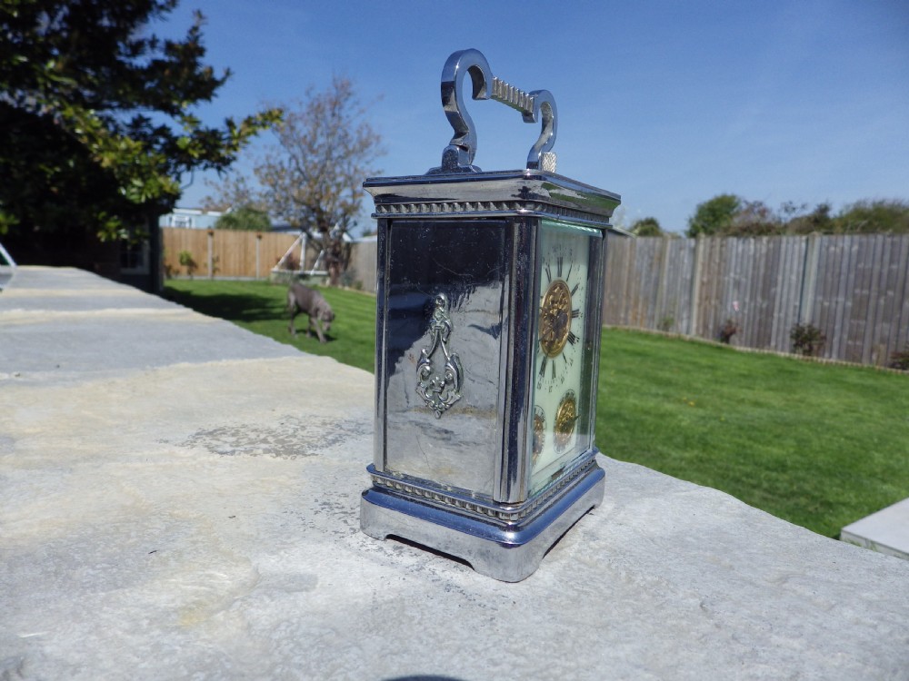 A silver-plated French carriage clock featuring a top carrying handle, a main white enamel dial with Roman numerals, and two subsidiary calendar dials set within a rectangular glass-panelled case. View IV
