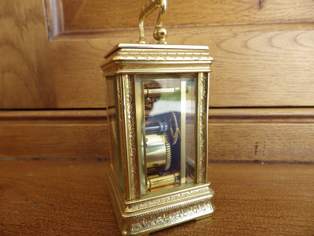 A miniature gilt brass carriage clock featuring detailed floral engraving, a folding top handle, glass side panels, and a gold-tone circular dial with Arabic numerals. View II