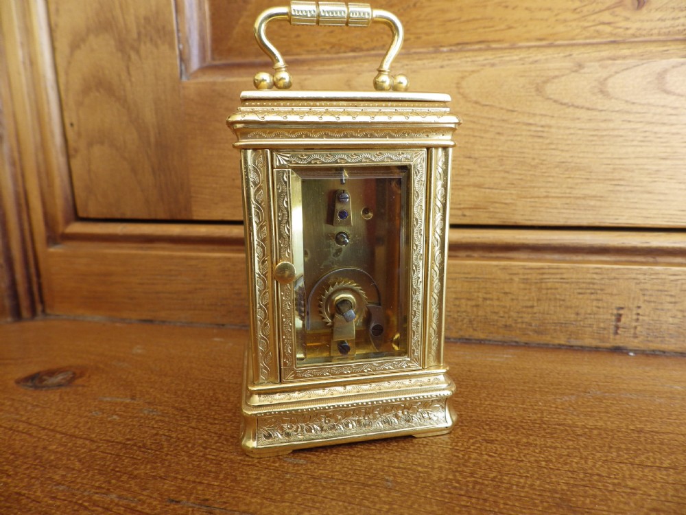 A miniature gilt brass carriage clock featuring detailed floral engraving, a folding top handle, glass side panels, and a gold-tone circular dial with Arabic numerals. View III