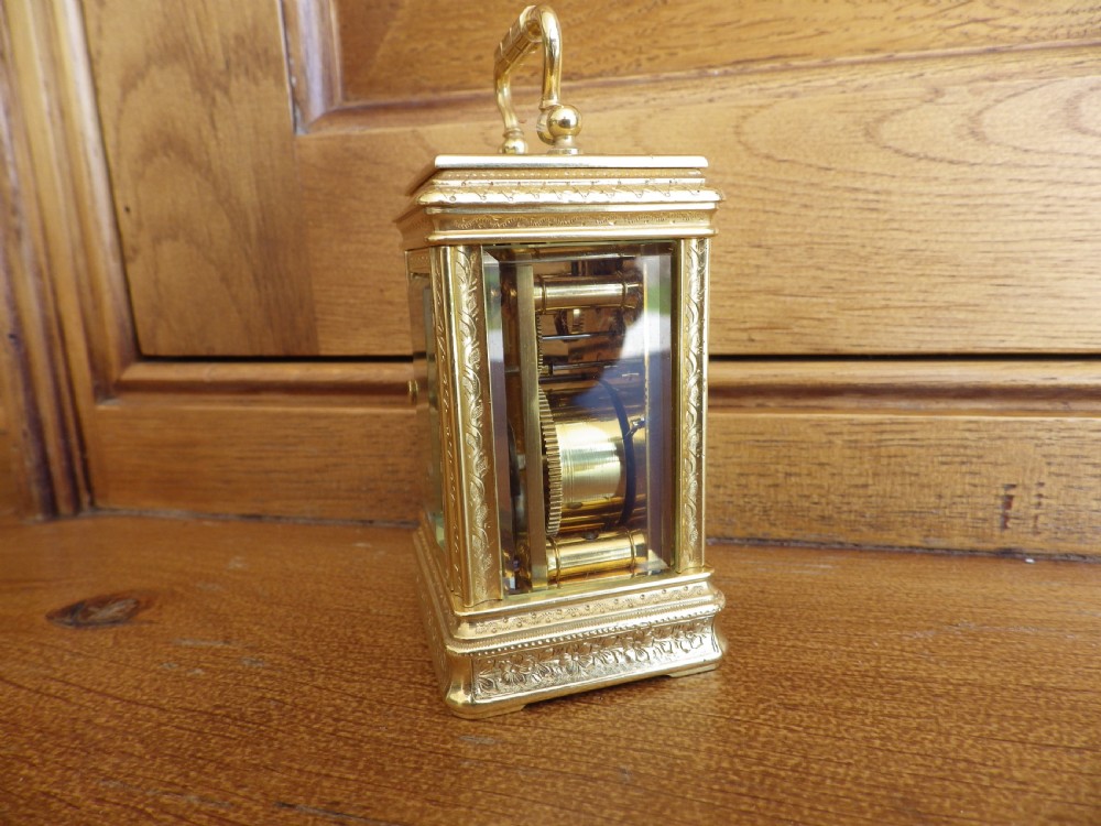 A miniature gilt brass carriage clock featuring detailed floral engraving, a folding top handle, glass side panels, and a gold-tone circular dial with Arabic numerals. View IV