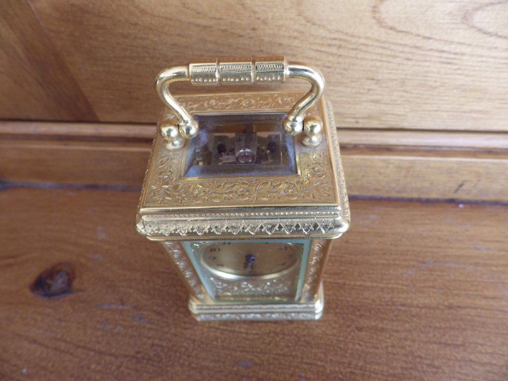 A miniature gilt brass carriage clock featuring detailed floral engraving, a folding top handle, glass side panels, and a gold-tone circular dial with Arabic numerals. View V