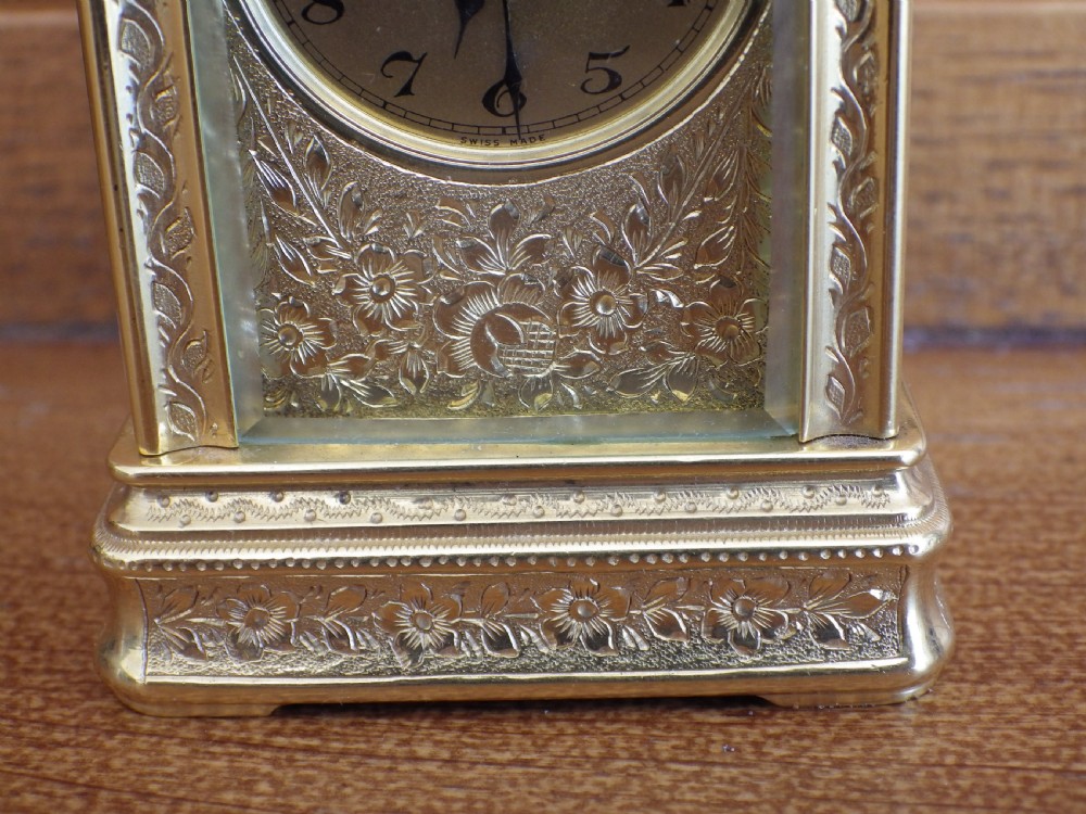 A miniature gilt brass carriage clock featuring detailed floral engraving, a folding top handle, glass side panels, and a gold-tone circular dial with Arabic numerals. View VI