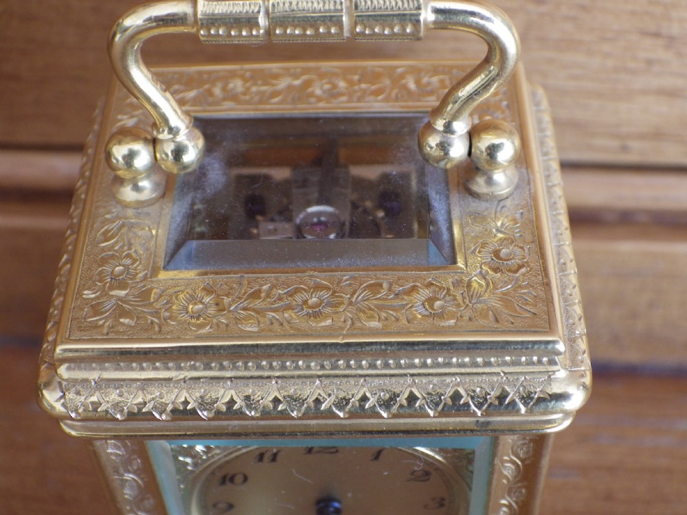 A miniature gilt brass carriage clock featuring detailed floral engraving, a folding top handle, glass side panels, and a gold-tone circular dial with Arabic numerals. View VII
