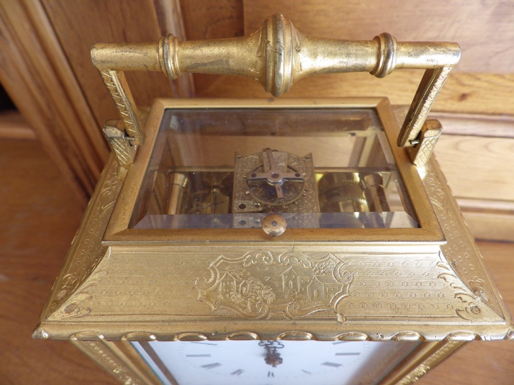 An antique gilt brass carriage clock featuring bevelled glass side panels, a top handle, a white enamel main dial with Roman numerals, and three subsidiary calendar dials below. View V