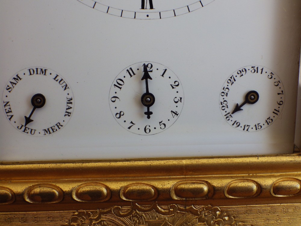 An antique gilt brass carriage clock featuring bevelled glass side panels, a top handle, a white enamel main dial with Roman numerals, and three subsidiary calendar dials below. View VII