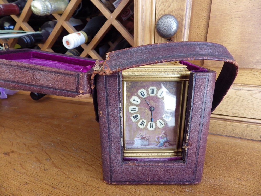 A rectangular gilt brass carriage clock with a hinged handle, featuring a painted enamel dial with Roman numeral cartouches and a scene depicting a figure seated in a garden setting. View IV