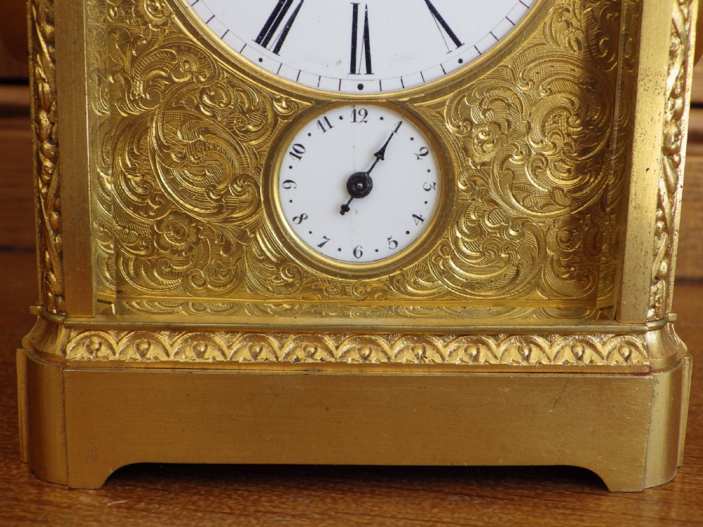 A French gilt brass carriage clock with an ornate foliate engraved face panel, white enamel dial with Roman numerals, secondary sub-dial, and decorative top handle. View V