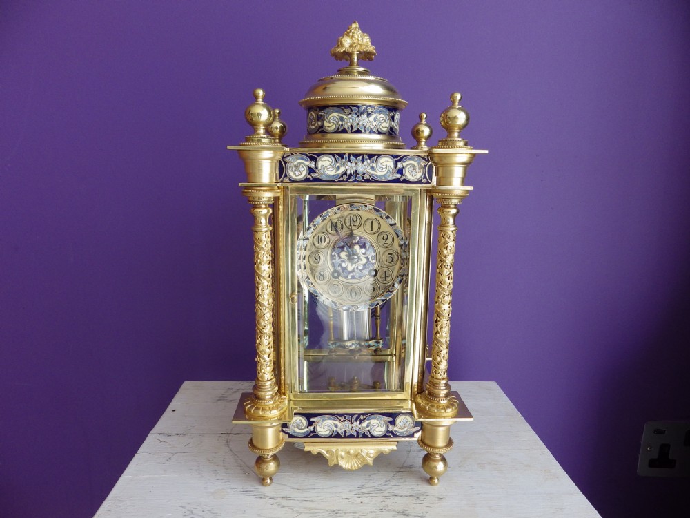 An antique French gilt brass crystal regulator clock featuring four clear glass sides, blue champleve enamel scrollwork, decorative corner columns, and an intricate dial.