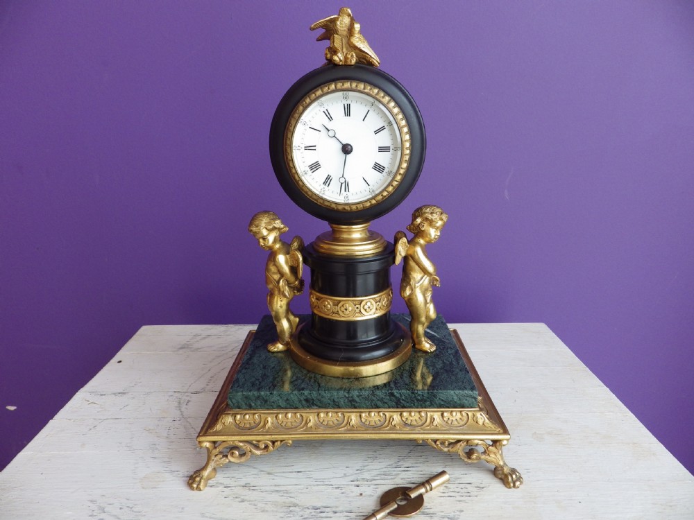 An 1880s French mantel clock featuring two gilt winged cherubs beside a central black column with a white enamel dial, set upon a green marble plinth with gilt feet.