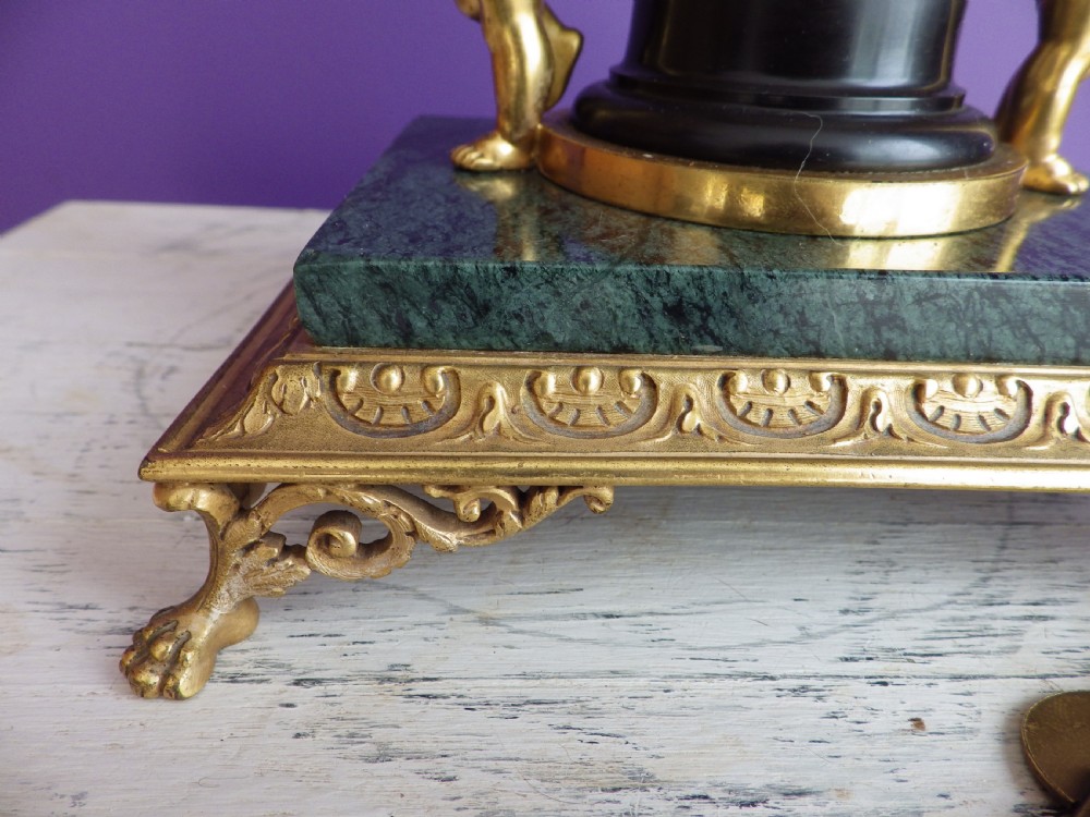 An 1880s French mantel clock featuring two gilt winged cherubs beside a central black column with a white enamel dial, set upon a green marble plinth with gilt feet. View V