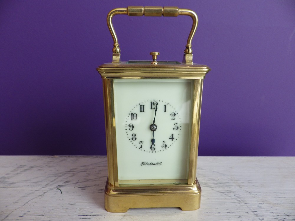 A brass French carriage clock featuring clear glass side panels, a white enamel dial with black numerals, and a hinged brass carrying handle.