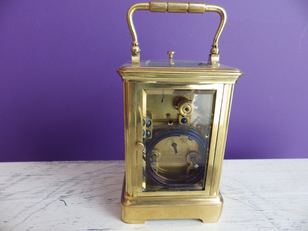 A brass French carriage clock featuring clear glass side panels, a white enamel dial with black numerals, and a hinged brass carrying handle. View III
