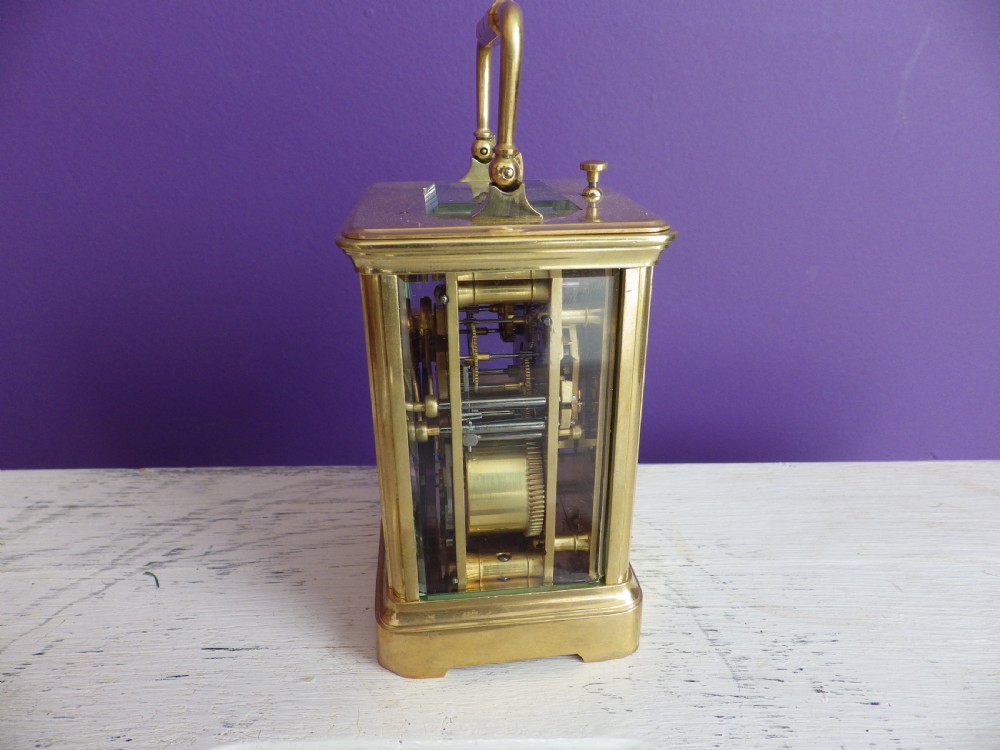 A brass French carriage clock featuring clear glass side panels, a white enamel dial with black numerals, and a hinged brass carrying handle. View IV