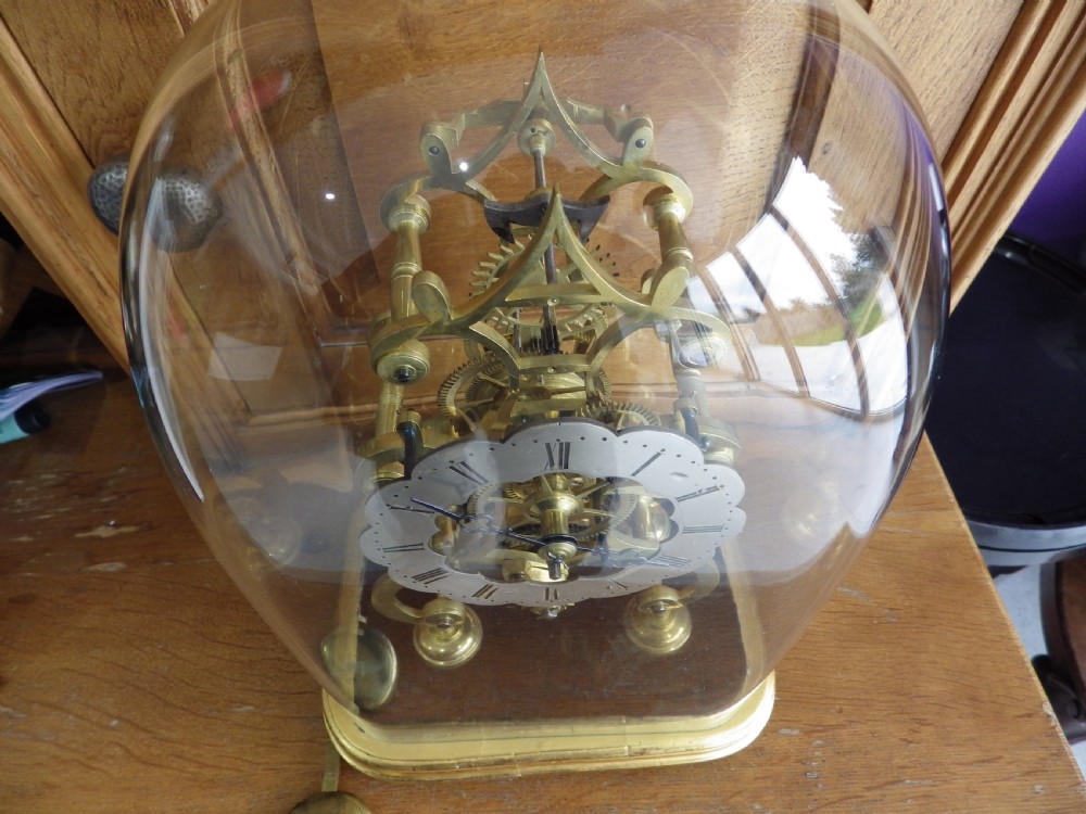 An 1870s English brass skeleton clock with an exposed mechanical movement and silvered chapter ring beneath a tall glass dome on a wooden base. View III