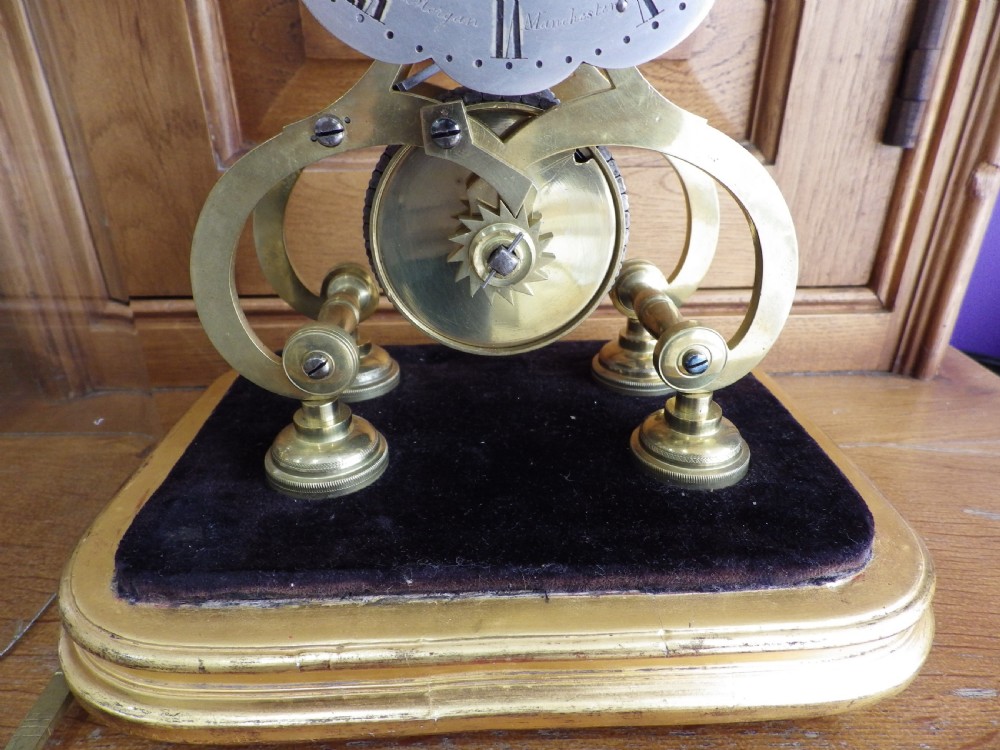 An 1870s English brass skeleton clock with an exposed mechanical movement and silvered chapter ring beneath a tall glass dome on a wooden base. View IV