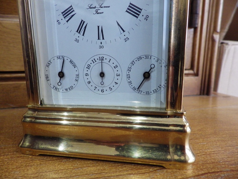 A brass carriage clock with bevelled glass panels, a white enamel dial, three subsidiary dials, and a top carrying handle resting on a dark box. View VII