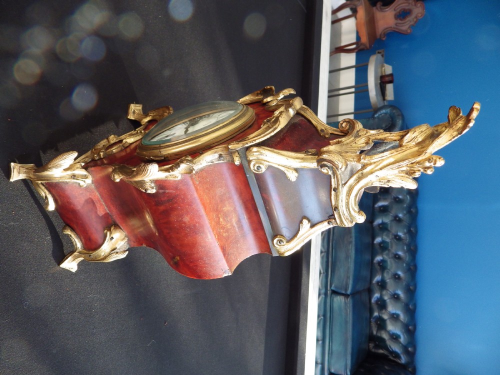 A late nineteenth-century French Rococo style mantel clock featuring ornate gilt mounts, a red tortoiseshell effect case, and a white enamel dial with Roman numerals. View II