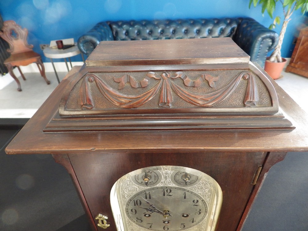 A dark wood 1930s bracket clock featuring a carved swag pediment, brass keyhole escutcheon, and an arched silvered dial with secondary chime sub-dials. View II