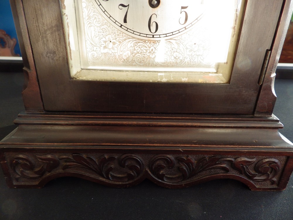A dark wood 1930s bracket clock featuring a carved swag pediment, brass keyhole escutcheon, and an arched silvered dial with secondary chime sub-dials. View III
