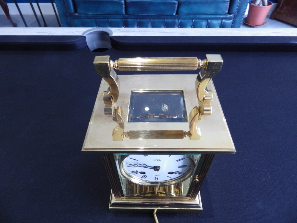A rectangular brass carriage clock with glass panels, a white dial featuring Roman numerals, an upper carrying handle, and a matching winding key. View II
