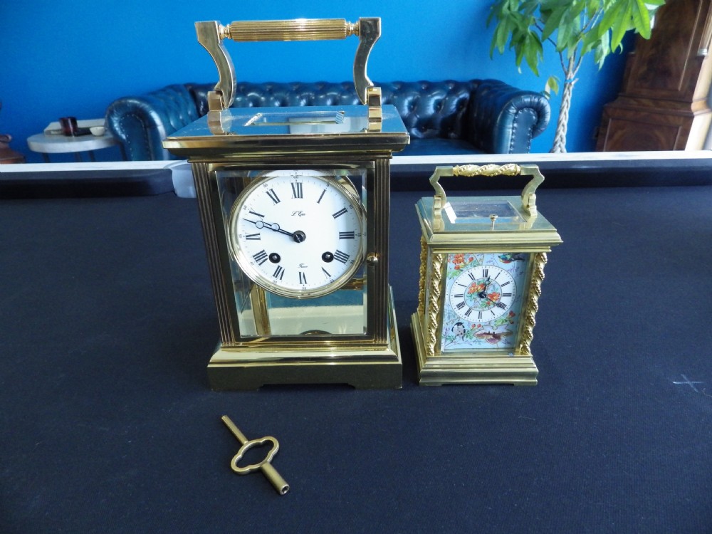 A rectangular brass carriage clock with glass panels, a white dial featuring Roman numerals, an upper carrying handle, and a matching winding key. View VII