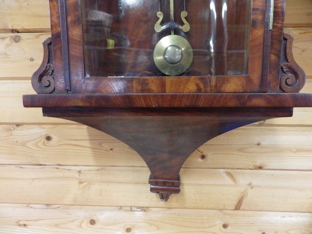 A Victorian flame mahogany wall clock featuring an arched top, painted dial with Roman numerals, carved fretwork, and a glass door displaying a brass pendulum. View II
