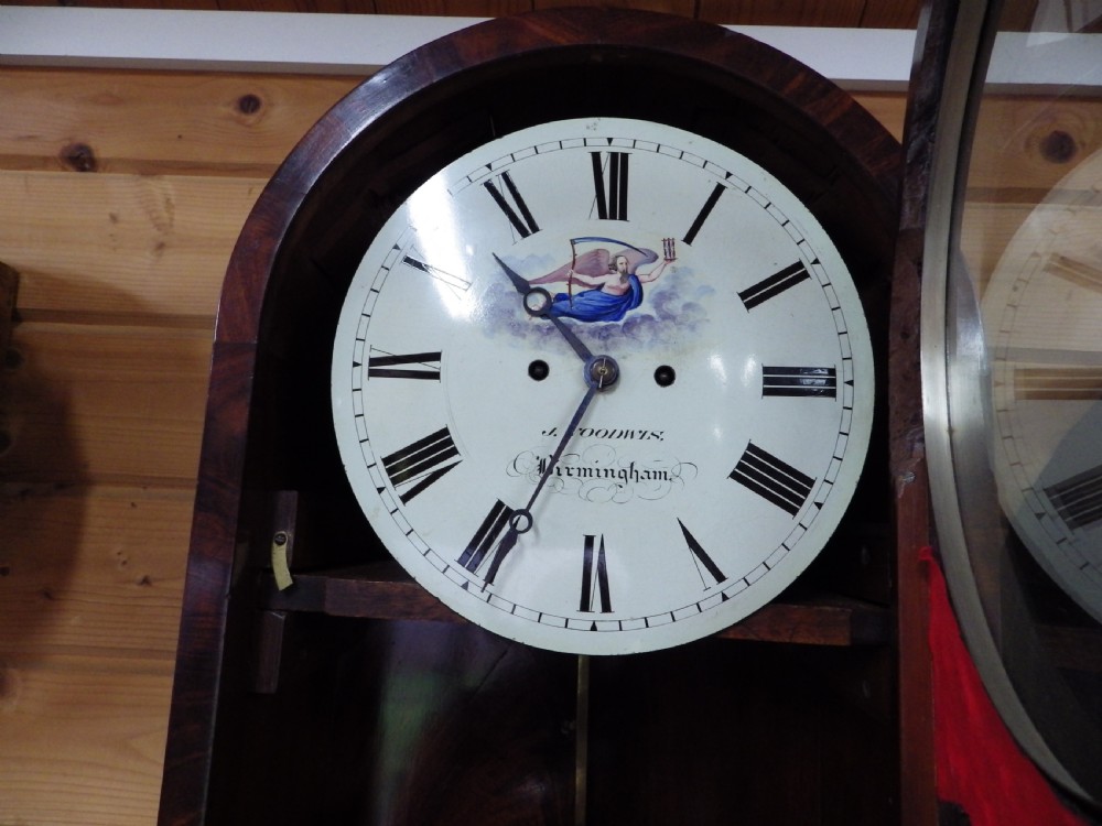 A Victorian flame mahogany wall clock featuring an arched top, painted dial with Roman numerals, carved fretwork, and a glass door displaying a brass pendulum. View III