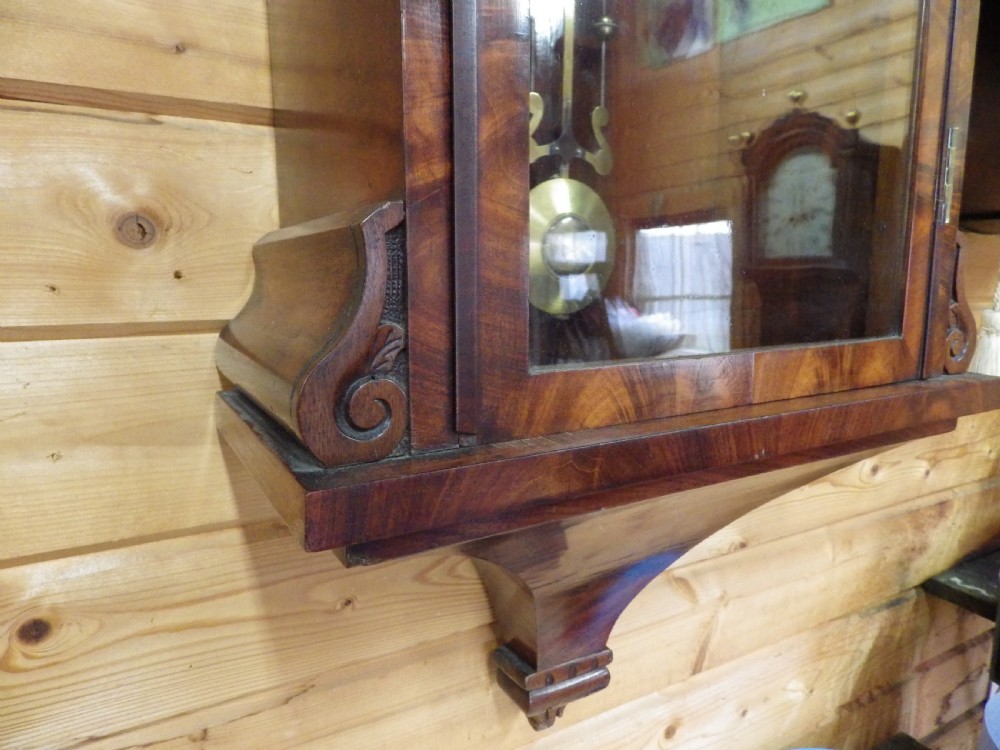 A Victorian flame mahogany wall clock featuring an arched top, painted dial with Roman numerals, carved fretwork, and a glass door displaying a brass pendulum. View VII