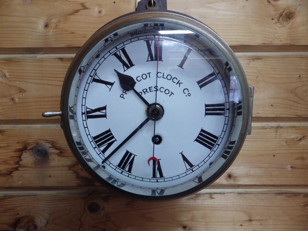 A circular Victorian brass ships clock featuring a white enamel dial with black Roman numerals, black spade hands, and a side hinge closure.