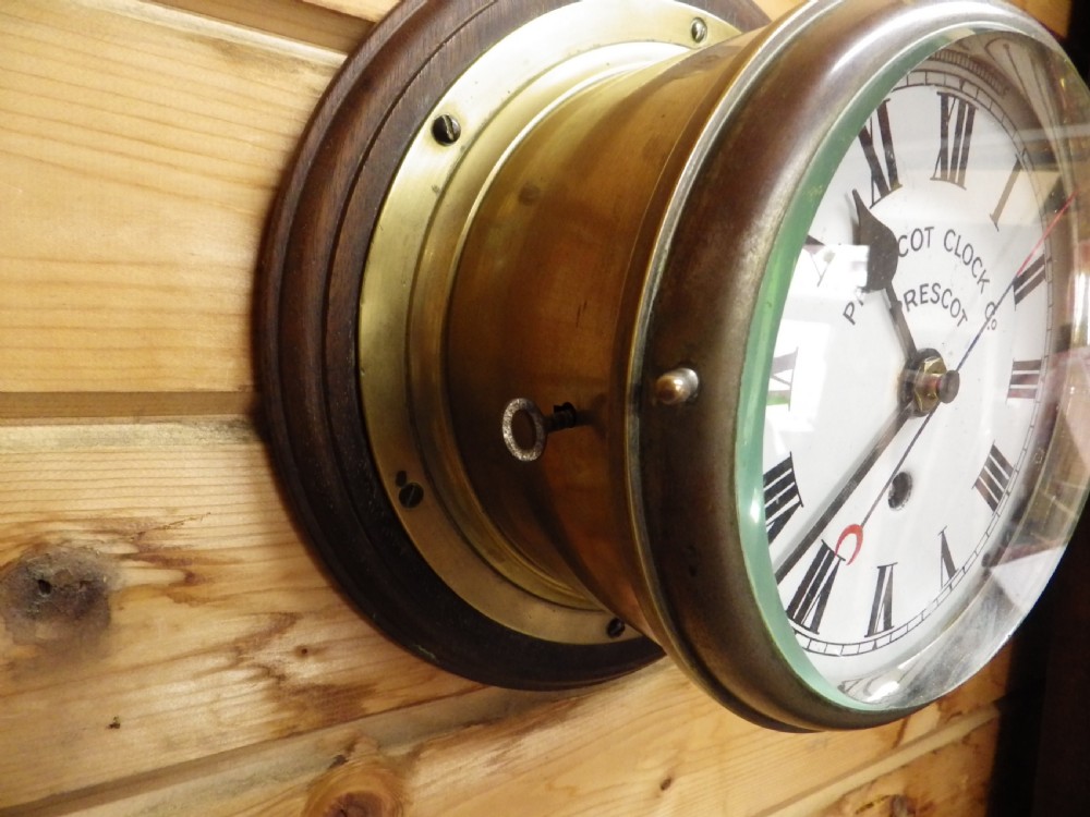 A circular Victorian brass ships clock featuring a white enamel dial with black Roman numerals, black spade hands, and a side hinge closure. View II