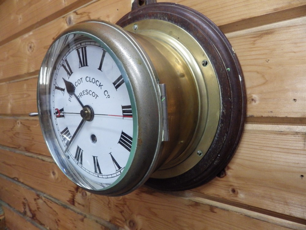 A circular Victorian brass ships clock featuring a white enamel dial with black Roman numerals, black spade hands, and a side hinge closure. View III