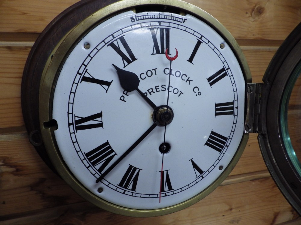 A circular Victorian brass ships clock featuring a white enamel dial with black Roman numerals, black spade hands, and a side hinge closure. View IV