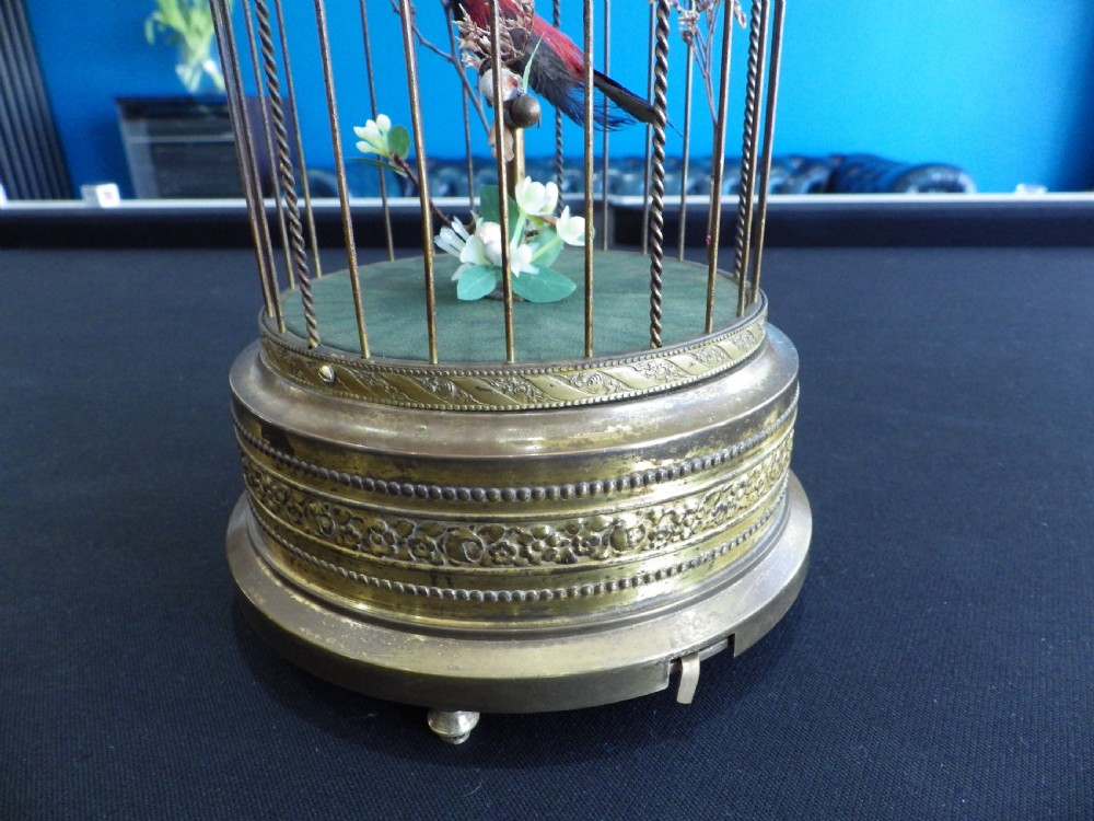A cylindrical brass domed cage containing a red and black feathered mechanical bird on a green fabric base with a top suspension loop. View II