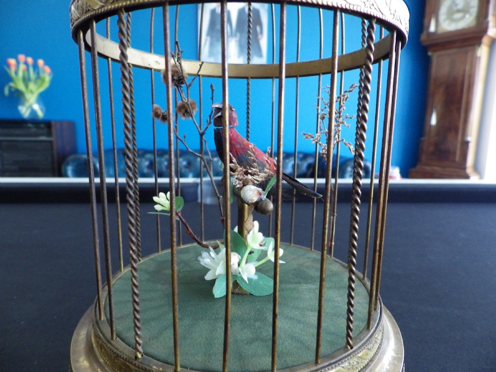 A cylindrical brass domed cage containing a red and black feathered mechanical bird on a green fabric base with a top suspension loop. View III