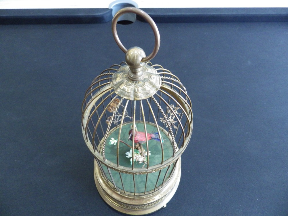A cylindrical brass domed cage containing a red and black feathered mechanical bird on a green fabric base with a top suspension loop. View IV