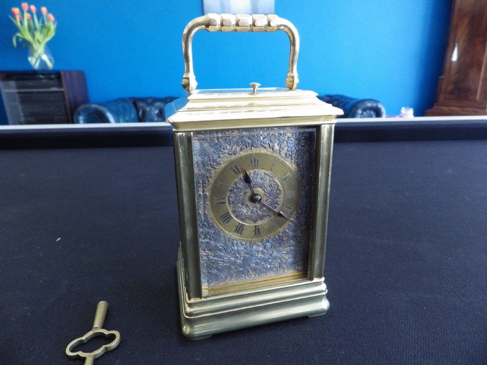 An antique brass carriage clock featuring an ornate silvered floral dial plate, Roman numerals, a top carrying handle, and an accompanying brass winding key.