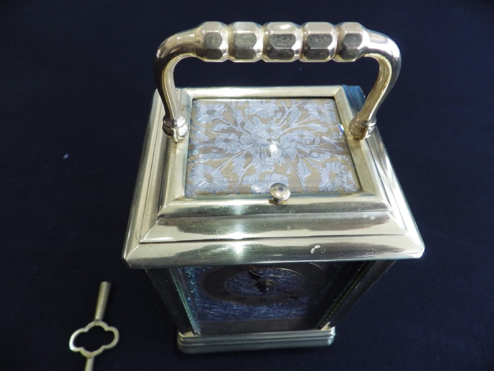 An antique brass carriage clock featuring an ornate silvered floral dial plate, Roman numerals, a top carrying handle, and an accompanying brass winding key. View II
