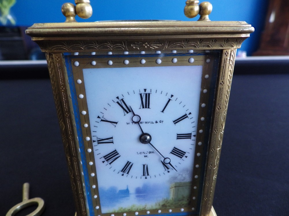 An engraved brass French carriage clock featuring a carrying handle, white enamel Roman numeral dial, and a hand-painted landscape panel. View II