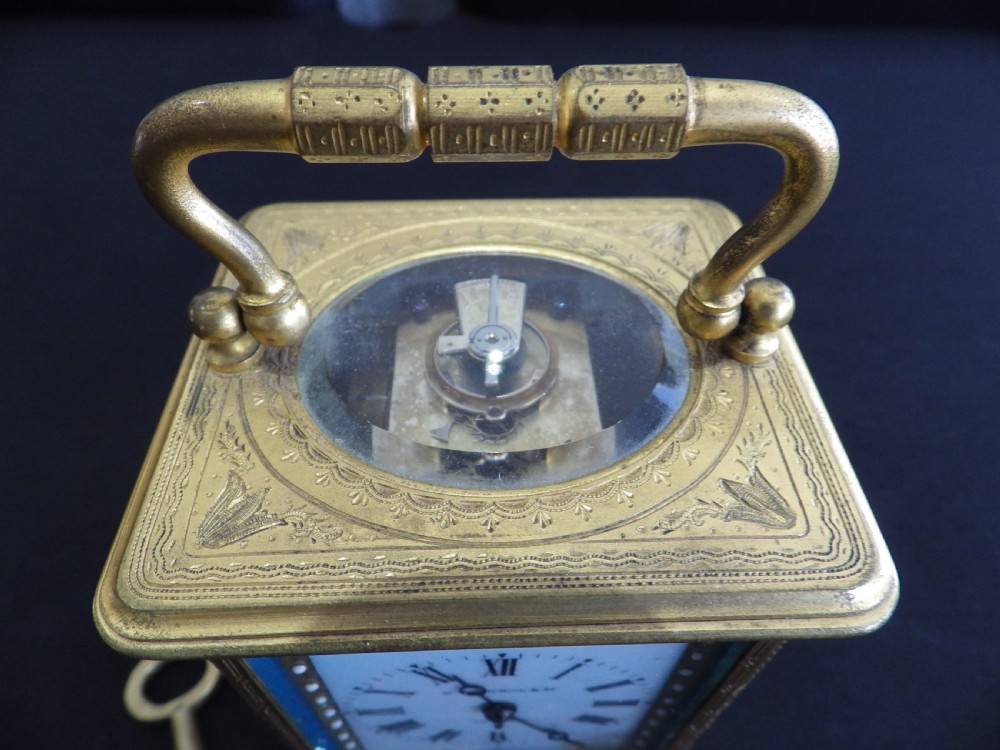 An engraved brass French carriage clock featuring a carrying handle, white enamel Roman numeral dial, and a hand-painted landscape panel. View IV