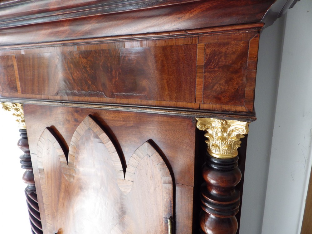 An antique flame mahogany longcase clock featuring a painted dial, swan-neck pediment with brass finials, and spirally turned column detailing. View VI