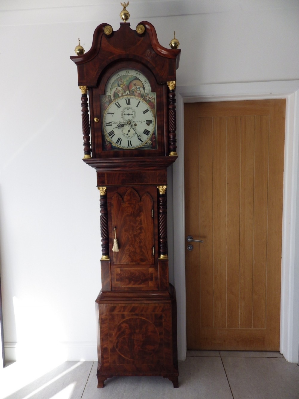 An antique flame mahogany longcase clock featuring a painted dial, swan-neck pediment with brass finials, and spirally turned column detailing.