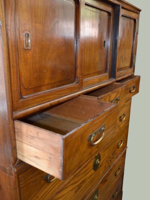 fig 1. A tall Taisho period Japanese elm tansu chest featuring prominent wood grain, circular brass escutcheons, sliding top cabinet doors, and multiple drawers.
