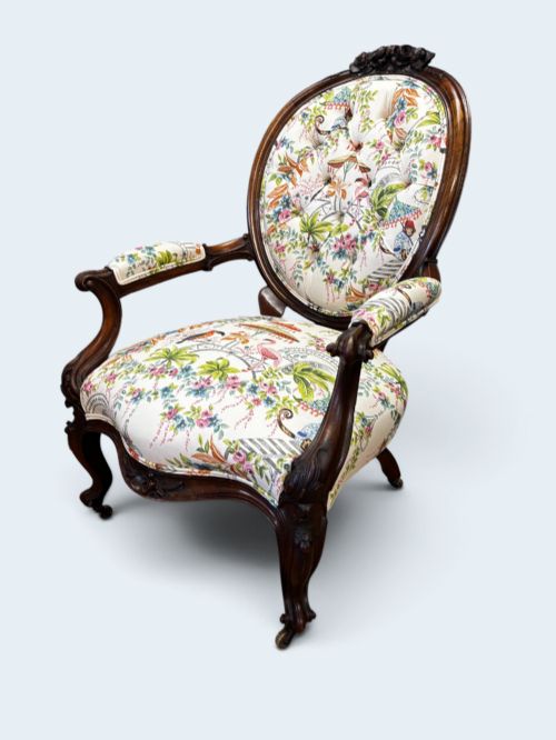 fig 1. A Victorian carved mahogany armchair featuring an oval button-backed backrest, padded armrests, and botanical floral upholstery set on cabriole legs with brass castors.